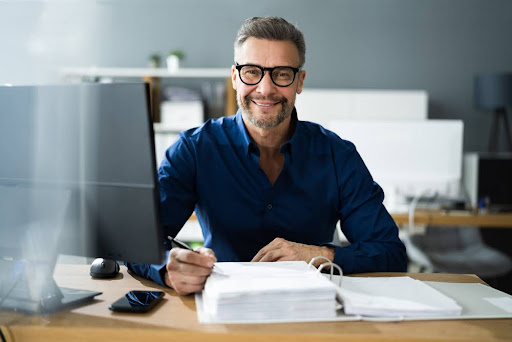 Person sits at desk smiling with stack of papers.